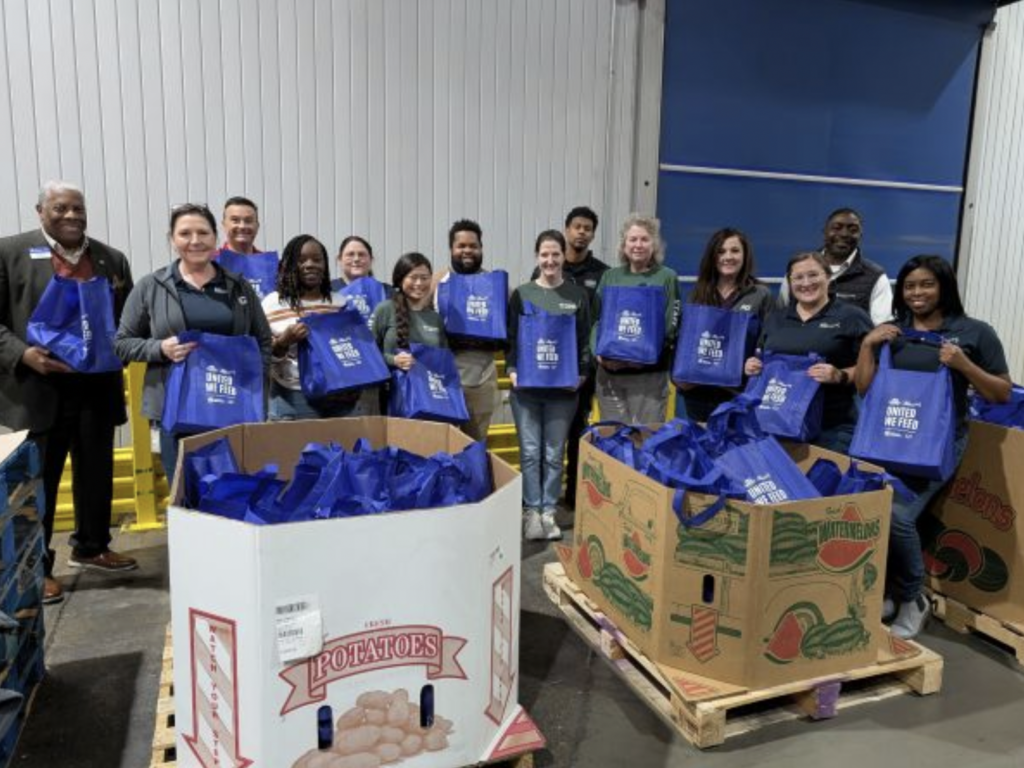 A group of about a dozen volunteers stand with bags in front of bins at an event to help empower women. 