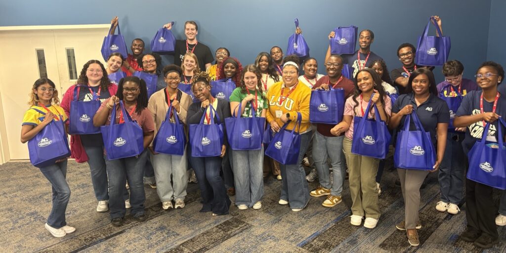Roeshawn Peters and Teach For America corps members smiling while holding up blue Associated Grocers reusable gift bags during summer teacher training.