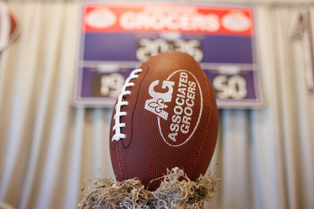 Brown football with Associated Grocers branding displayed on a decorative stand, positioned in front of an out-of-focus scoreboard showing a tied score of 50–50.